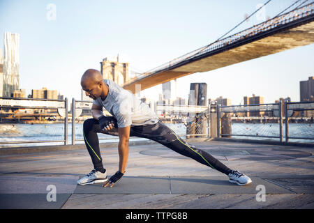 Volle Länge des männlichen Athleten trainieren auf der Promenade mit Brooklyn Bridge im Hintergrund Stockfoto