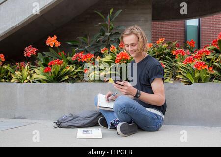 Männliche Kursteilnehmer sitzen von Blumen mit Büchern, an Sms lächelnd Stockfoto
