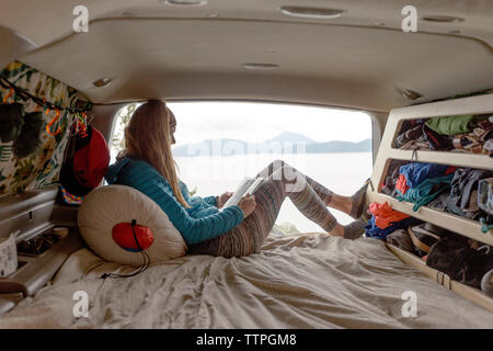 Seitenansicht der Frau mit Buch beim Sitzen in Camper trailer gegen See Stockfoto