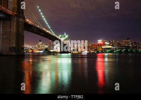 Beleuchtete Brooklyn Brücke über den Fluss gegen die beleuchtete Stadt Stockfoto