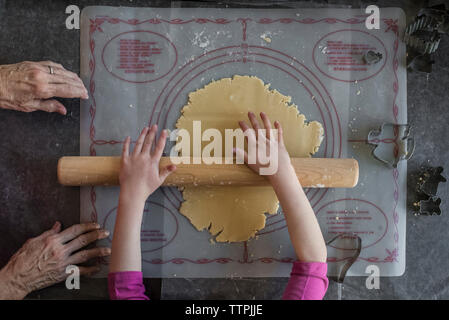 7/8 Hände der Enkelin rolling Plätzchenteig von Großmutter auf den Tisch zu Hause. Stockfoto