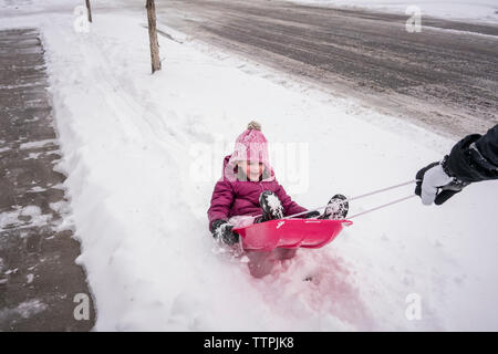 7/8 Hand des Vaters ziehen Tochter sitzen auf dem Schlitten auf der schneebedeckten Straße Stockfoto