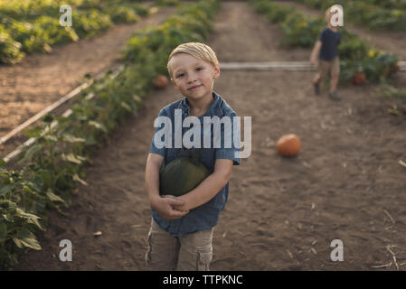 Hohen winkel Portrait von Junge mit Kürbis mit Bruder, der im Hintergrund auf der Farm Stockfoto