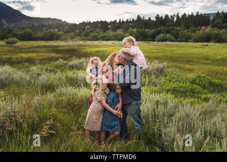 Romantische Eltern küssen beim Stehen mit Töchtern auf der Wiese gegen Himmel im Wald Stockfoto