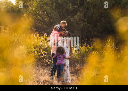Romantische Eltern küssen beim Stehen mit Töchtern auf Feld gegen Bäume im Park Stockfoto