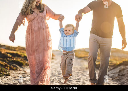 Glückliche Eltern Holding's Sohn Hände beim Gehen auf Sand am Strand gegen den klaren Himmel bei Sonnenuntergang Stockfoto