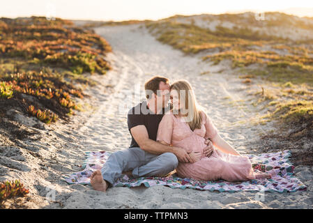 Freundlich lächelnde Paare sitzen auf Decke am Strand gegen den klaren Himmel bei Sonnenuntergang Stockfoto