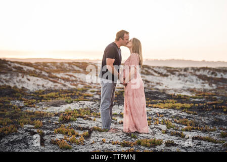 Seitenansicht des Romantisches Paar küssen beim Stehen am Strand gegen den klaren Himmel bei Sonnenuntergang Stockfoto