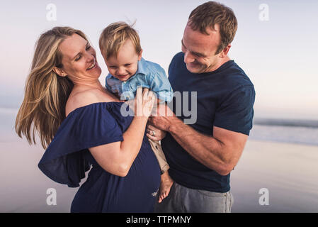 Glückliche Eltern, die niedlichen Sohn beim Stehen am Strand gegen Himmel bei Sonnenuntergang Stockfoto