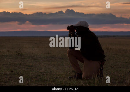 Die ganze Länge der Mann mit Fernglas während kniend auf Feld in der Serengeti National Park bei Sonnenuntergang Stockfoto