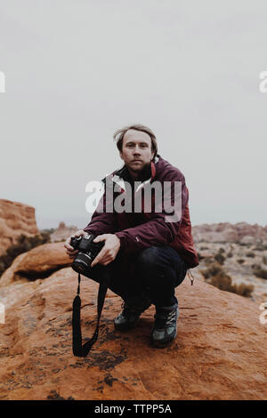 Porträt der Mann hält Kamera beim kauern auf Rock gegen Sky Stockfoto