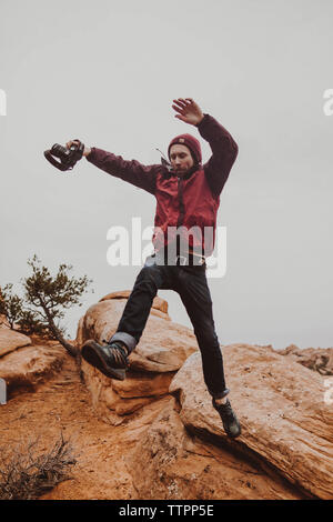 Low Angle View der Mann hält Kamera beim Springen auf den Felsen gegen Sky Stockfoto