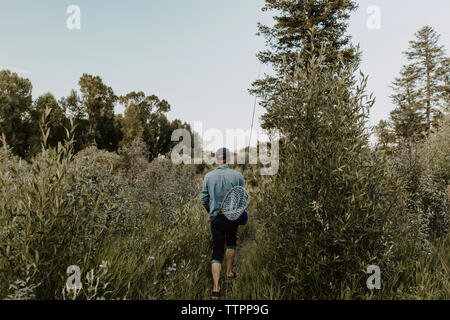 Ansicht der Rückseite des Mann mit angelausrüstung Wandern inmitten von Pflanzen gegen klaren Himmel im Wald Stockfoto