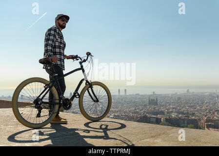 Seitenansicht der Mann mit dem Fahrrad stehend auf stützmauer über Stadtbild gegen Himmel während der sonnigen Tag Stockfoto