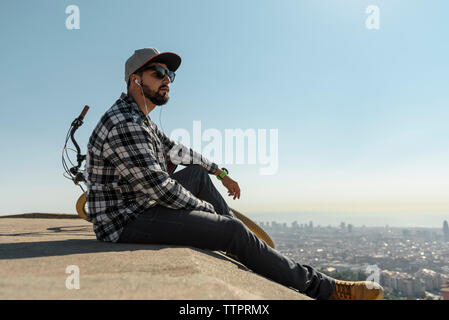 Seitenansicht des Menschen tragen Sonnenbrillen Musik hören beim Sitzen auf der Mauer gegen den klaren blauen Himmel während der sonnigen Tag Stockfoto