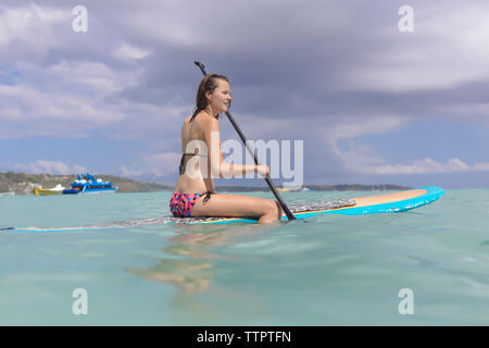 Frau im Bikini paddleboarding am Meer gegen bewölkter Himmel Stockfoto