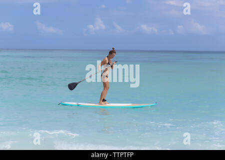 Frau im Bikini paddleboarding am Meer gegen Himmel während der sonnigen Tag Stockfoto