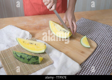 7/8 Hände von Frau schneiden Wassermelone auf dem Tisch zu Hause Stockfoto