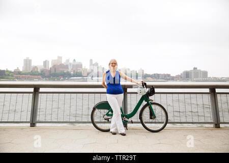 Portrait von glücklich, reife Frau, die mit dem Fahrrad auf der Promenade in Stadt gegen den klaren Himmel Stockfoto