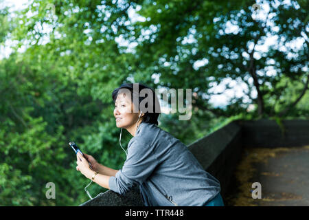 Nachdenkliche Frau Musik hören während lehnte sich auf Stützmauer gegen Filialen Stockfoto