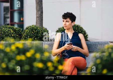 Nachdenkliche Frau Musik hören beim Sitzen auf Stützmauer gegen Gebäude Stockfoto