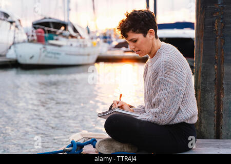 Seitenansicht der Frau schriftlich beim Sitzen auf Pier am Hafen gegen Himmel bei Sonnenuntergang Stockfoto