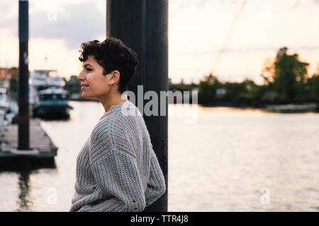 Seitenansicht der lächelnden nachdenkliche Frau weg, während Sie am Hafen gegen Himmel stehen bei Sonnenuntergang Stockfoto
