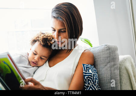 Low Angle View von Mutter Lesung Buch für Sohn beim Sitzen auf dem Sofa zu Hause gegen Fenster Stockfoto