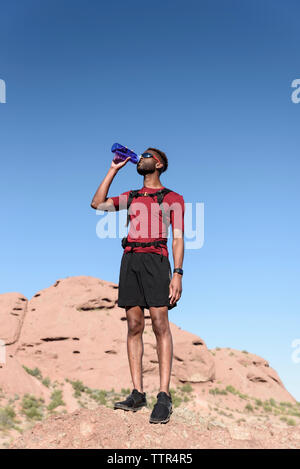 Low Angle View männlicher Wanderer Trinkwasser beim Stehen auf Rock Formation gegen den klaren blauen Himmel Stockfoto