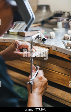 Zugeschnittenes Bild der weiblichen Handwerker mit handsäge während Herstellung von Schmuck auf hölzernen Tisch in Werkstatt Stockfoto