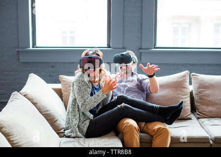Glückliche Mitarbeiter tragen Virtual Reality Simulatoren beim Sitzen auf einem Sofa im Büro Stockfoto