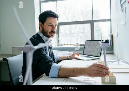 Geschäftsmann, der ein Notebook benutzt, während er das Modell einer Windkraftanlage auf dem Schreibtisch arrangiert Stockfoto