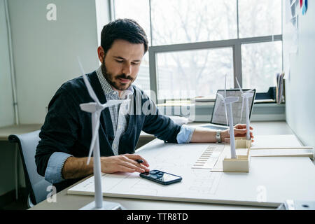 Geschäftsmann, der sein Smartphone benutzt, während er Windkraftanlagenmodelle auf dem Schreibtisch arrangiert Stockfoto