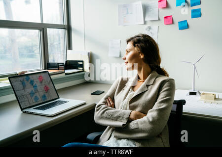 Geschäftsfrau mit überkreuzten Armen, die auf einem Stuhl am Schreibtisch im Büro sitzt Stockfoto