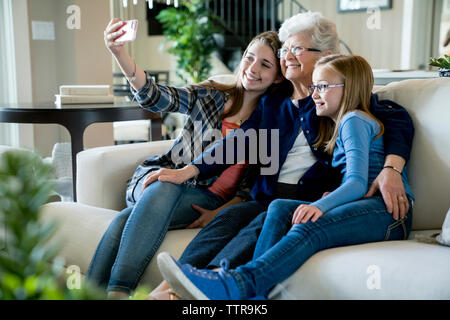 Gerne Großmutter und Enkelinnen unter selfie durch Smart Phone beim Sitzen auf dem Sofa Stockfoto