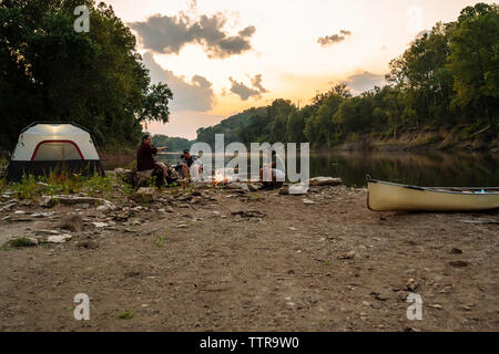 Männliche Freunde sprechen beim Sitzen auf einem Campingplatz gegen Himmel bei Sonnenuntergang Stockfoto
