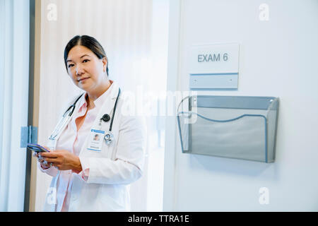 Portrait von zuversichtlich Ärztin mittels Smart Phone im Stehen gegen die Tür im Krankenhaus Stockfoto