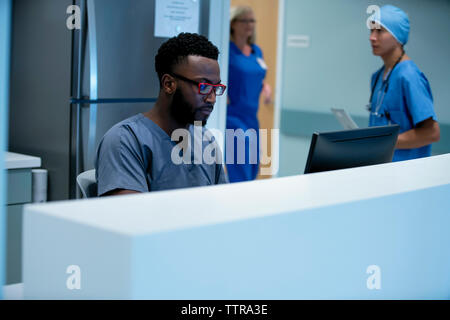 Zuversichtlich Arzt mit dem Desktop Computer am Krankenhaus Rezeption mit Kollegen im Hintergrund arbeiten Stockfoto