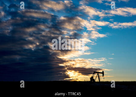 Mitte der Abstand der Silhouette pumpjack am Ölindustrie über Feld gegen bewölkter Himmel bei Sonnenuntergang Stockfoto