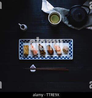 Überblick über verschiedene Sushi in der Platte mit Stäbchen und grüner Tee auf schwarz Tabelle angeordnet Stockfoto