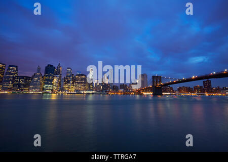 Blick auf das beleuchtete Skyline und Brooklyn Bridge gegen bewölkter Himmel bei Nacht Stockfoto