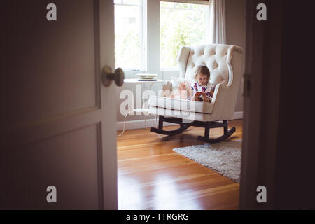 Mädchen lesen Bild Buch sitzend auf Schaukelstuhl zu Hause durch Tür gesehen Stockfoto