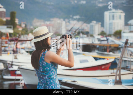 Seitenansicht der Frau mit Hut fotografieren Boote am Hafen mit Kamera beim Stehen in der Stadt Stockfoto