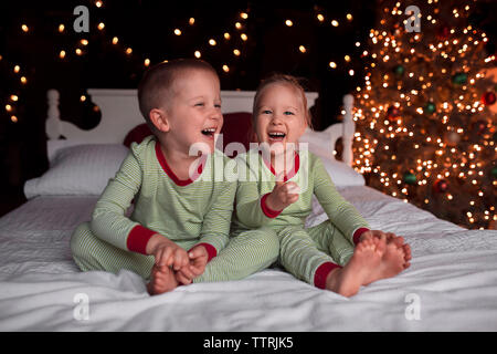 Fröhliche Geschwister sitzen zu Hause vor dem beleuchteten weihnachtsbaum auf dem Bett Stockfoto