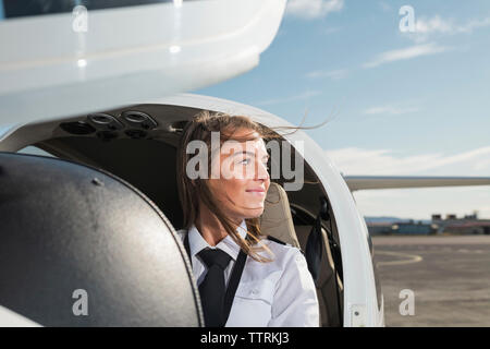 Lächelnd nachdenklich Pilotin weg schauen beim Sitzen im Flugzeug gegen Himmel am Flughafen Stockfoto