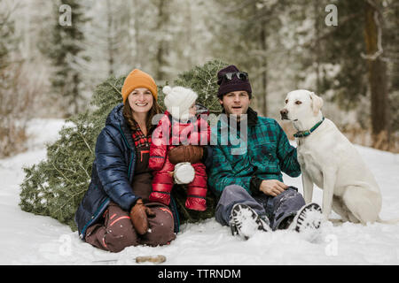 Portrait von Eltern mit Tochter und Hund sitzt von Pine Tree auf Schnee im Wald bedeckt Stockfoto