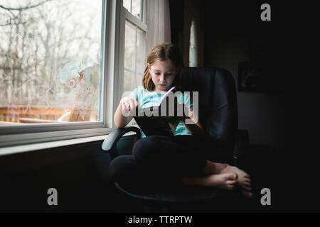 Volle Länge des Mädchen studieren beim Sitzen auf Stuhl von Fenster zu Hause Stockfoto