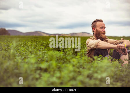 Gerne männliche Hipster sitzt inmitten von Pflanzen auf dem Feld gegen Sky Stockfoto