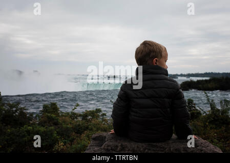 Ansicht der Rückseite des Junge sitzt auf Felsen sitzend gegen Niagara Falls Stockfoto