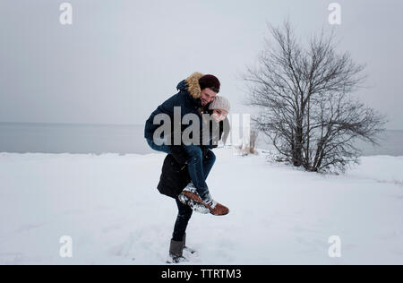 Glückliche Frau huckepack männlichen Freund beim Stehen auf Schnee Strand gegen Himmel Stockfoto
